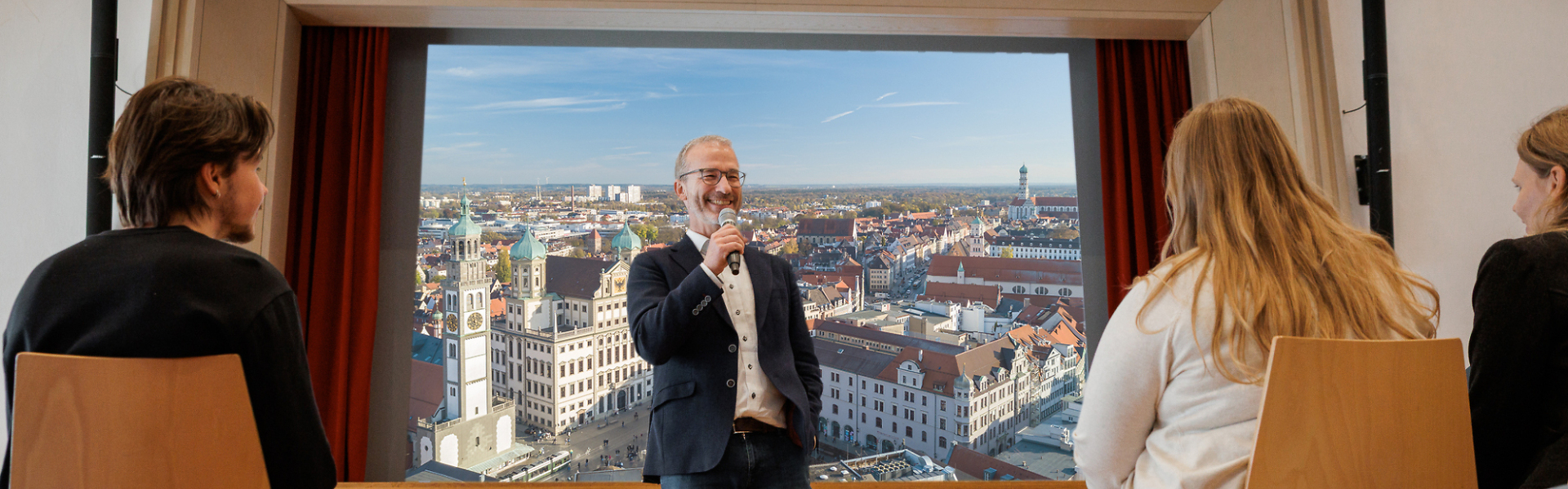 Person steht mit Mikrofon vor einer großen Leinwand mit Stadtansicht und spricht zu sitzenden Teilnehmenden in einem Veranstaltungsraum.