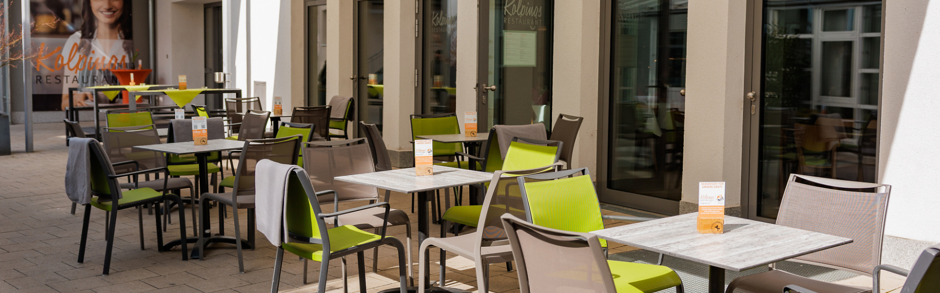 Außenbereich eines Restaurants mit mehreren gedeckten Tischen und Stühlen entlang der Fassade unter einer Markise.