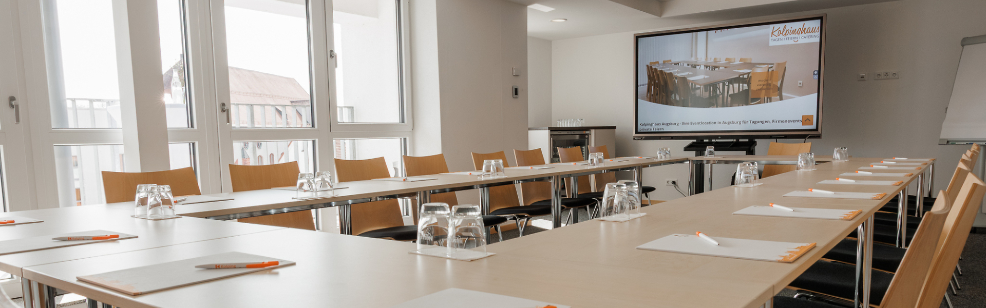 Heller Seminarraum Domblick mit U-förmig angeordneten Tischen, vorbereitet mit Notizblöcken, Stiften und Gläsern sowie einem Bildschirm für Präsentationen im Hintergrund.