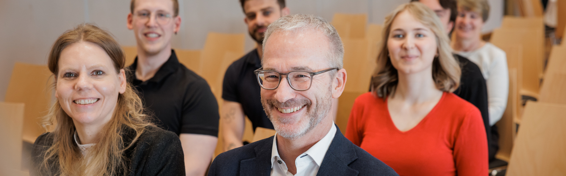 „Mehrere Teilnehmende sitzen in Reihen in einem Veranstaltungsraum und schauen lächelnd nach vorne, im Vordergrund ist ein Mikrofon zu sehen.