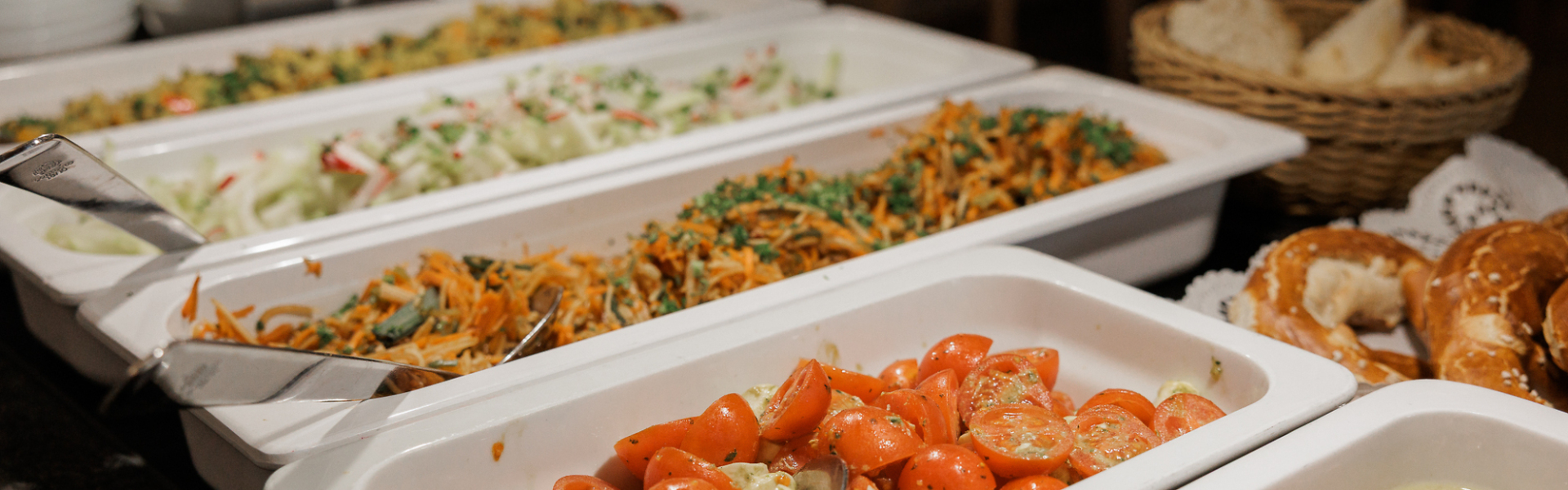 Buffet mit verschiedenen Salaten und Beilagen in weißen Schalen sowie Brotkorb im Hintergrund.