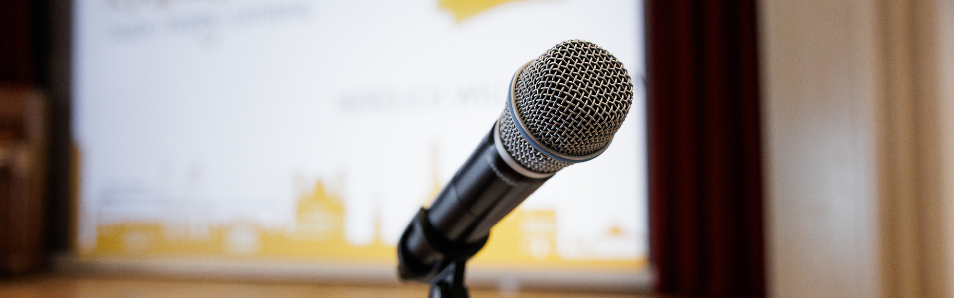 Mikrofon auf einem Stativ vor einer Bühne mit Präsentationsleinwand im Hintergrund.