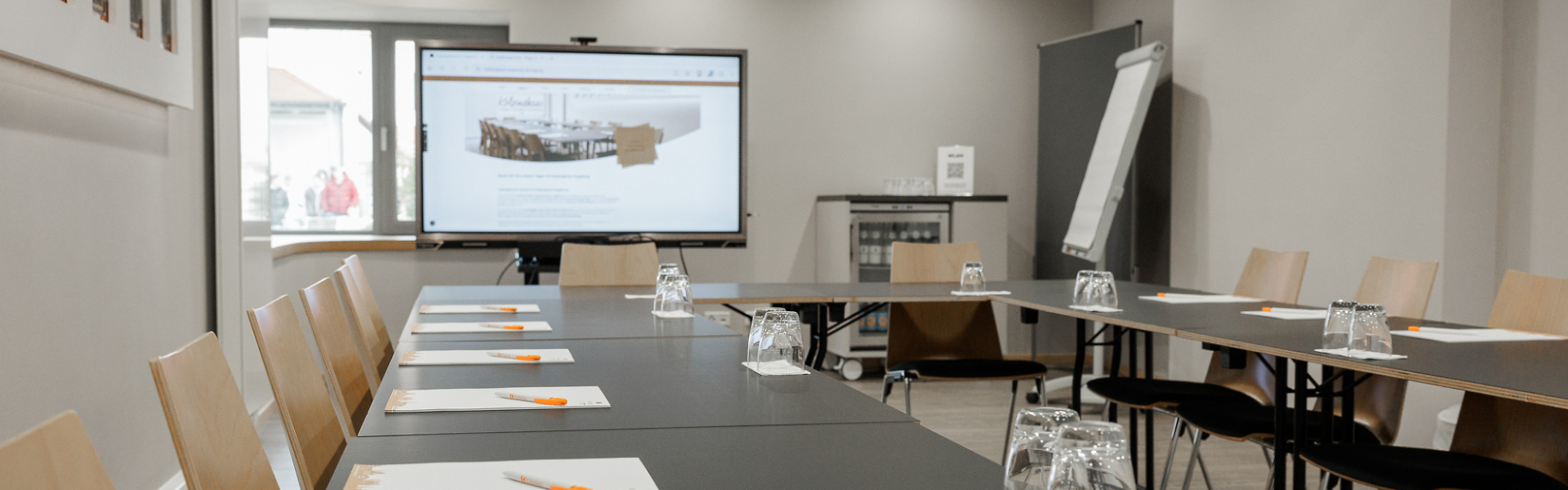 Moderner Seminarraum mit U-förmig angeordneten Tischen, ausgestattet mit Notizblöcken, Stiften und Gläsern sowie Bildschirm und Flipchart im Hintergrund.