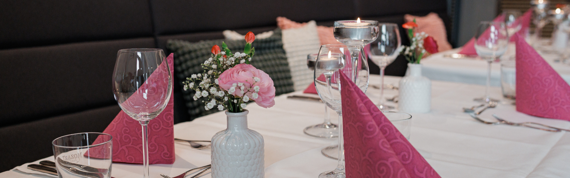 Ein festlich gedeckter Restauranttisch mit weißen Tischdecken, gefalteten rosafarbenen Servietten, Gläsern, Besteck und kleinen Blumenvasen als Dekoration.
