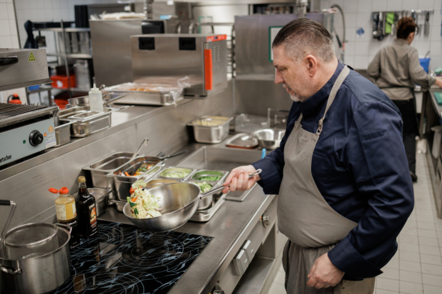 Koch steht in einer Großküche am Herd und bereitet in einer Pfanne ein Gericht mit Gemüse zu.