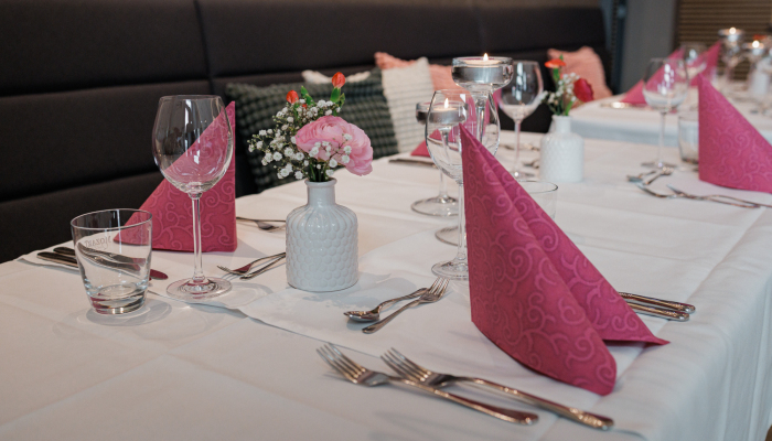 Ein festlich gedeckter Restauranttisch mit weißen Tischdecken, gefalteten rosafarbenen Servietten, Gläsern, Besteck und kleinen Blumenvasen als Dekoration.