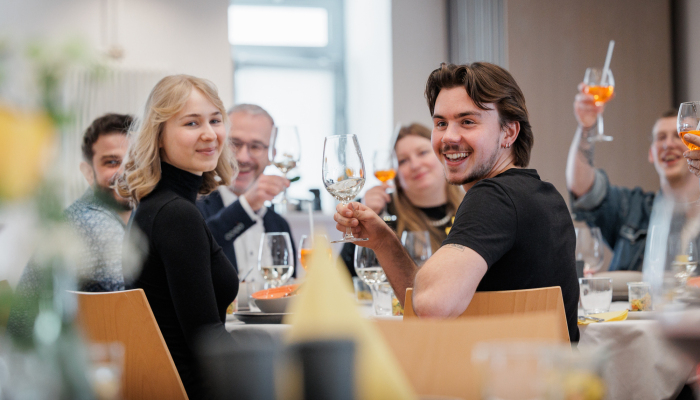 Mehrere Personen sitzen gemeinsam an einem Tisch, stoßen mit Getränken an und lachen in einem Restaurant.