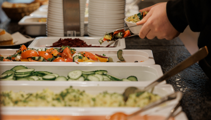 Person bedient sich an einem Buffet mit verschiedenen Salaten und Beilagen und legt Speisen auf einen Teller.