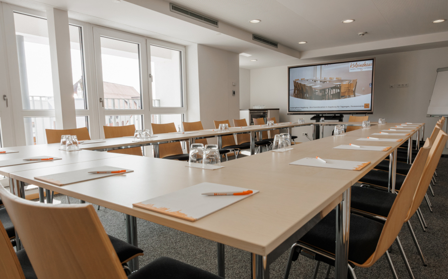 Heller Seminarraum Domblick mit U-förmig angeordneten Tischen, vorbereitet mit Notizblöcken, Stiften und Gläsern sowie einem Bildschirm für Präsentationen im Hintergrund.