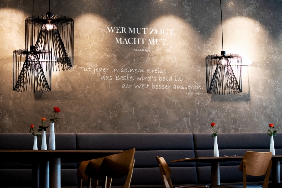 Stilvoller Restaurantbereich mit modernen Hängeleuchten und dekorativer Wand mit inspirierendem Zitat, gedeckte Tische mit einzelnen Blumenvasen.