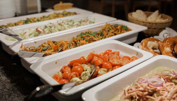 Buffet mit verschiedenen Salaten und Beilagen in weißen Schalen sowie Brotkorb im Hintergrund.