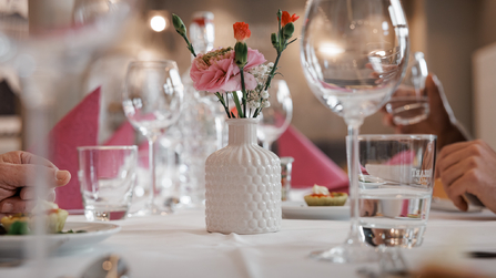 Gedeckter Tisch mit Gläsern, Blumenvasen und Servietten im Vordergrund.
