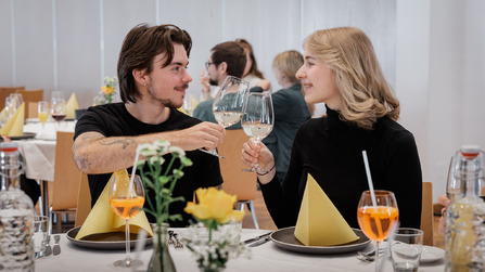 Zwei Personen sitzen im Restaurant, lächeln sich an und stoßen mit Weingläsern an.