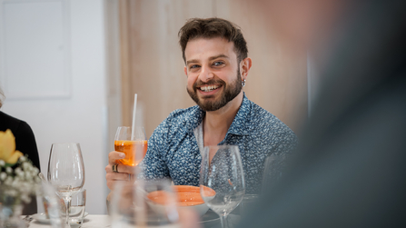 Ein Mann sitzt im Restaurant, lächelt in die Kamera und hält ein Getränk in der Hand.