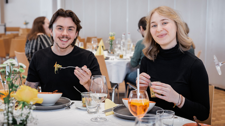 Zwei Personen sitzen am Tisch, essen und lächeln in die Kamera.