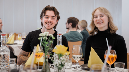 Zwei Personen sitzen nebeneinander am gedeckten Tisch und lächeln in die Kamera.