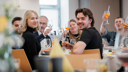 Eine Gruppe von Menschen sitzt im Restaurant, dreht sich zur Kamera und stößt gemeinsam mit Getränken an.