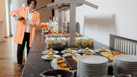 Ein Buffet mit verschiedenen kleinen Speisen, Desserts und Gläsern ist auf einer Theke angerichtet, während im Hintergrund eine Frau ein Getränk einschenkt.